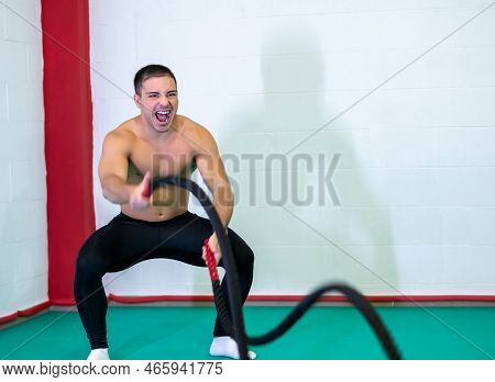 A Handsome Muscular Young Man Using Battle Ropes For Exercise In A Gym. He Focuses On Performing The