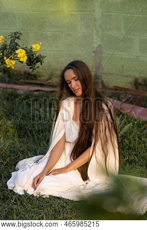 A Girl With Very Long Hair Sits On The Grass In A Torpic Landscape. White Dress Of The Bride. Health