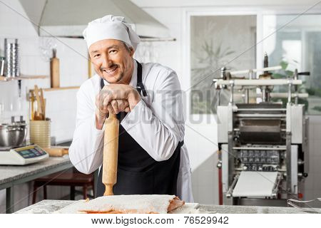 Portrait of happy male chef holding rolling pin at counter while preparing pasta in commercial kitchen