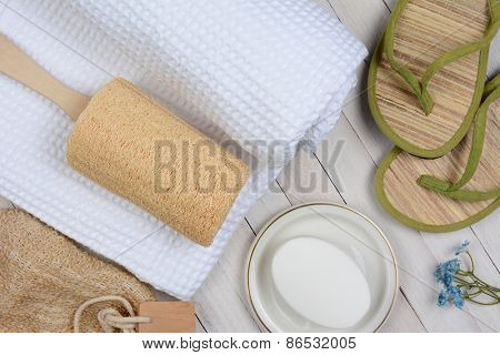 Spa and bath still life. High angle shot of bathroom accessories including, towel, soap, loofah and sandals, on a rustic white wood surface.