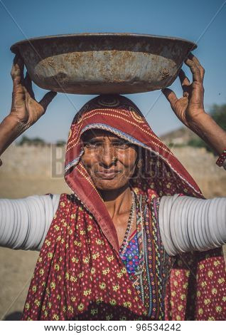 GODWAR REGION, INDIA - 14 FEBRUARY 2015: Rabari tribeswoman stands in field wearing saree and upper-arm bracelets. Balances bucket on head. Post-processed with grain, texture and colour effect.