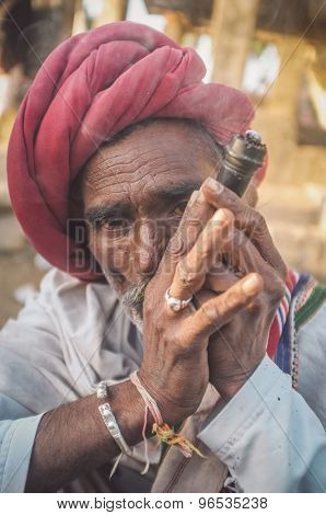 GODWAR REGION, INDIA - 14 FEBRUARY 2015: Elderly Rabari tribesman with red turban and blanket smokes chillum. Post-processed with grain, texture and colour effect.