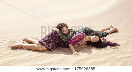 Desert women lying on sand outdoors. Dehydration, overheating, thirst and heat stroke concept image with two sisters outdoors in the nature. Two arabian girls lost in desert during journey.