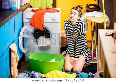 Young housewife sitting sadly near the washing machine with lots of colorful clothes on the floor at home