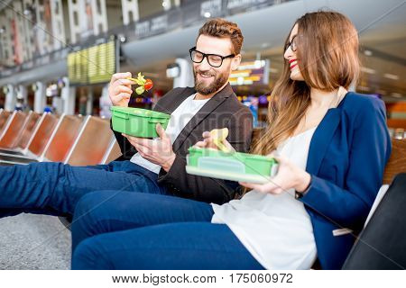 Elegant business couple eating with lunch boxes sitting at the waiting hall in the airport. Having a snack during business trip