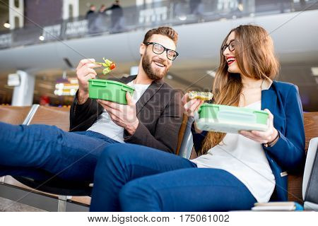 Elegant business couple eating with lunch boxes sitting at the waiting hall in the airport. Having a snack during business trip