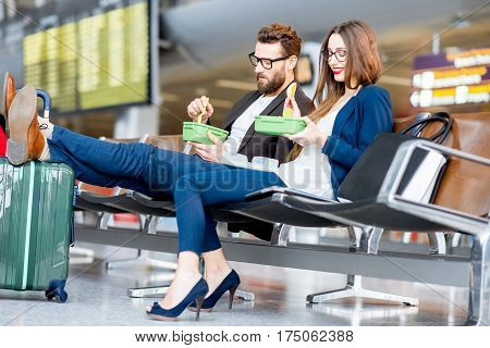 Elegant business couple eating with lunch boxes sitting at the waiting hall in the airport. Having a snack during business trip
