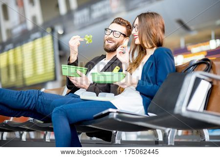 Elegant business couple eating with lunch boxes sitting at the waiting hall in the airport. Having a snack during business trip