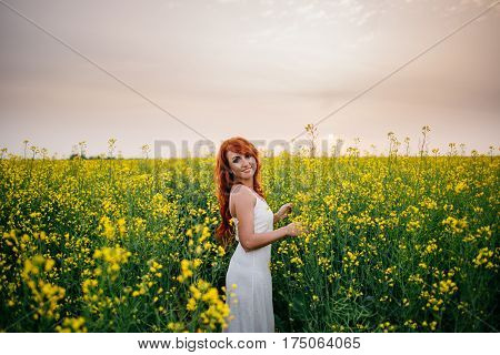 Young beautiful red-haired woman in a blooming rapeseed field at sunset