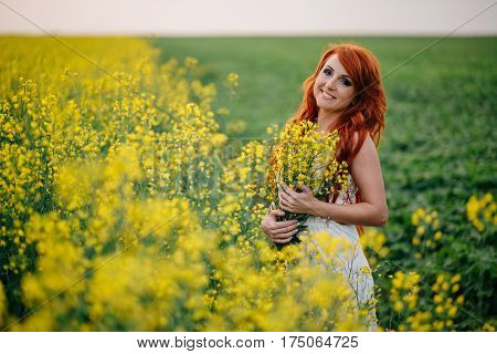 Young beautiful red-haired woman in a blooming rapeseed field at sunset