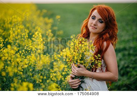 Young beautiful red-haired woman in a blooming rapeseed field at sunset