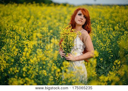 Young beautiful red-haired woman in a blooming rapeseed field at sunset