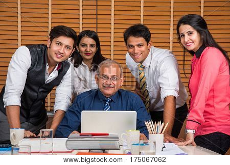 Satisfied with new plan. Group of four to five Indian cheerful young people and one experienced businessman looking at laptop with smile while leaning to the table in office