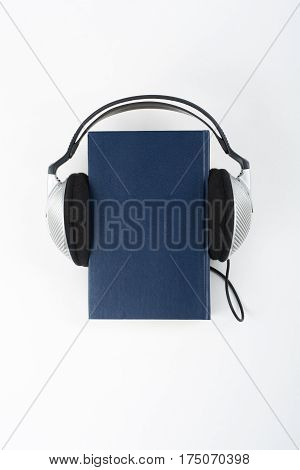 Audiobook on white background. Headphones put over blue hardback book empty cover copy space for ad text. Distance education e-learning concept