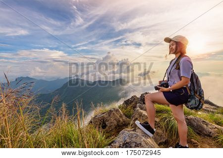 Hiker asian young woman happy with camera backpack and caps looking beautiful landscape nature of mountain and colourful sky at sunset on viewpoint Phu Chi Fa Forest Park Chiang Rai Thailand