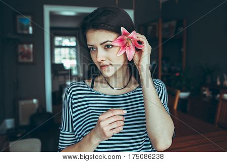 Closeup portrait of smiling pensive Caucasian young beautiful woman model with long hair in striped t-shirt holding large pink flower near ear hair looking away toned with filters