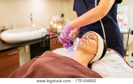 people, beauty, cosmetic treatment, cosmetology and technology concept - beautician with microdermabrasion device doing face exfoliation to young woman lying at spa parlor