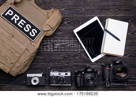 Bulletproof vest,retro and professional DSLR camera,notebook,pen and PC tablet on dark wooden table.Top view