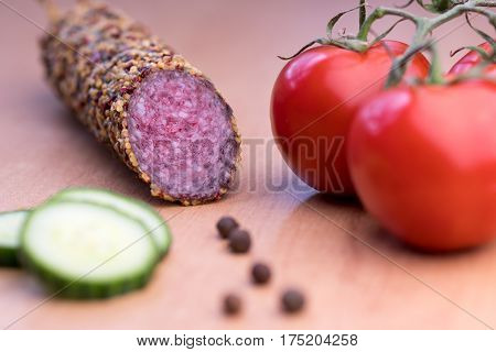 Delicious smoked sausage whole on wooden board with slices of cucumbers and tomatoes. View from above.
