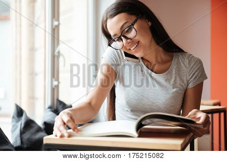 Photo of young caucasian woman indoors talking by mobile phone and reading magazine. Coworking concept.