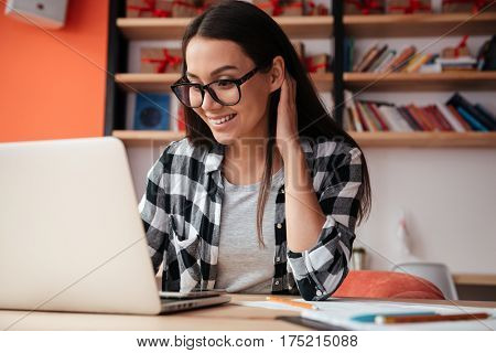 Photo of pretty young caucasian woman wearing glasses sitting indoors using laptop computer. Coworking concept.
