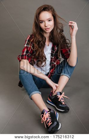Full length of attractive young woman in plaid shirt and jeand sitting over grey background