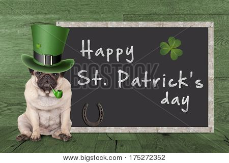 cute pug puppy dog with leprechaun hat for st. patrick's day smoking pipe sitting next blank chalkboard sign with horseshoe and shamrock on green wooden background