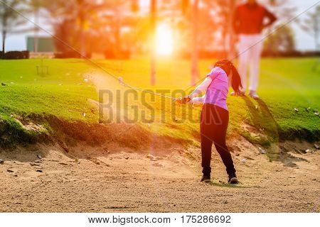 female golf player swinging golf from sand bunker during sunset with flare