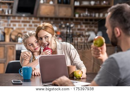 Happy young family in kitchen using laptop and eating fresh apples