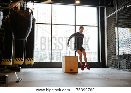 Athletic man which jumping from the box in gym with window on background