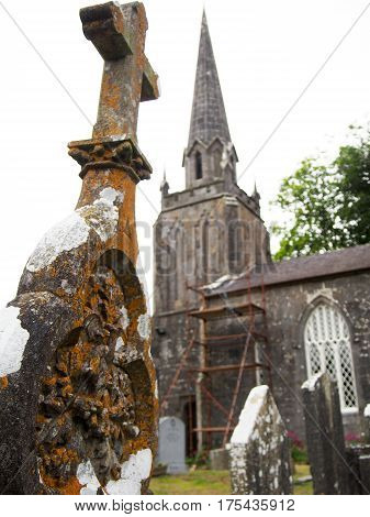 Detail of an old weathered headstone with a rural Catholic church in the background in Cork County Ireland.