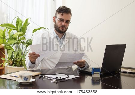 A doctor in a white coat in a doctor's office sits at a table and fills papers Doctor's office.