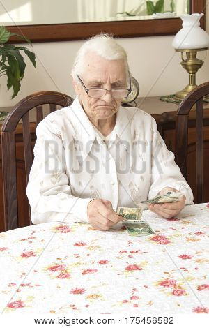 The old woman counting money while sitting at the table. 
Pension. Old woman counts the money. Old woman with a sad face counts money while sitting at the table.