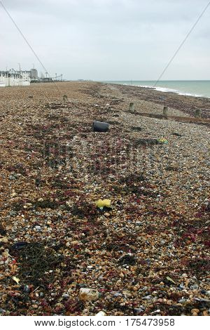 Strandline on Worthing beach in Sussex. Lots of litter mixed in amongst the seaweed. Beach with pebbles and breakwaters. Sea visible in the distance.