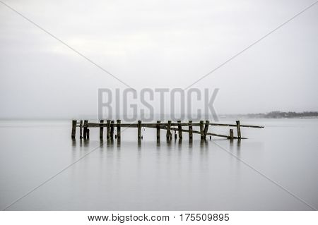 Broken pier in calm tranquil water posts reflecting with misty foggy background seascape