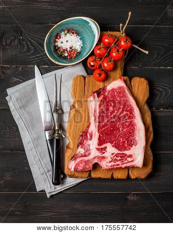Raw fresh meat Club Steak, seasoning and meat fork and knife on dark wooden background, top view