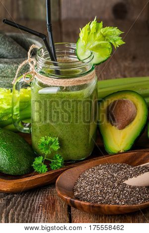 Healthy green juice smoothie with straw surrounded by avocado, cucumber,  celery and chia seeds on wooden background