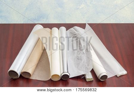 Several rolls of the plastic oven bags plastic food wrap aluminum foil and various parchment paper for household use on a dark red wooden surface