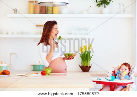 young pregnant mother and infant baby in highchair having lunch on kitchen at home
