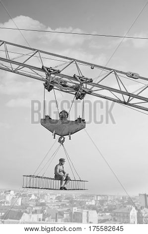 Away from city madness. Vertical monochrome shot of retro builder man relaxing having lunch sitting high above the city on a crossbar hanging from a construction crane worker industry business people