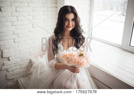 Luxury bride brunette with curly hair sit near the window. Girl with a bouquet
