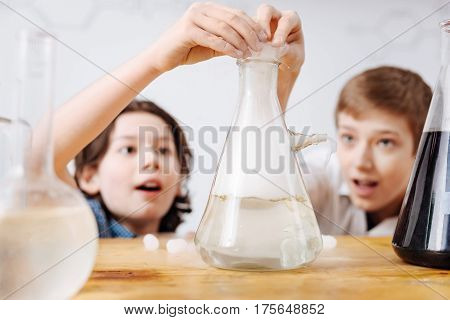 Science project. Intelligent clever exited boys hiding behind the table and putting some reagents into a flask while doing their science project