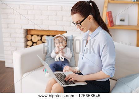 Modern motherhood. Inventive energetic strong woman analyzing the latest sales report using her laptop while her daughter sitting on a couch near her