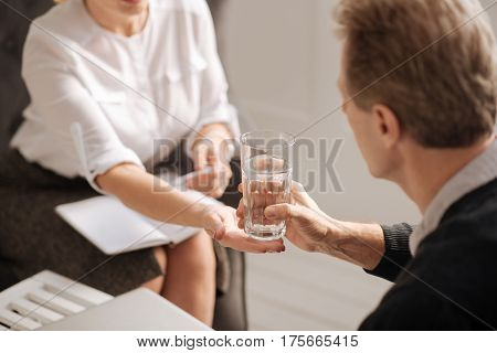 It is for you. Selective focus of a glass of water being held by a nice sympathetic pleasant therapist while being given to the patient