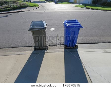 Two wheeled garbage containers (grey and blue) set out on street curbside on collection day, casting shadows on sidewalk.