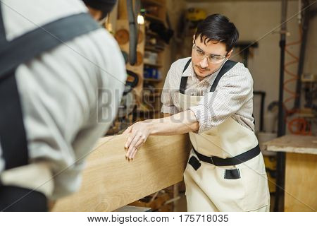 Two men holding timber on woodworking machine. Workplace equipped sleeves for hood. Rounded templates hanging on wall near machine-tool.