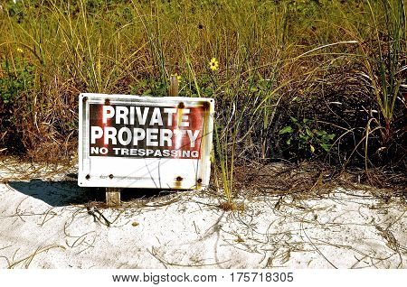 Private Property and NoTrespassing sign  in front of weeds, seed oats, and grasses on a sandy beach.