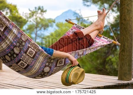 Person relaxing in Hummock holding Travel Hat in Hand at Summer Garden with Forest and Mountains on Background Landscape