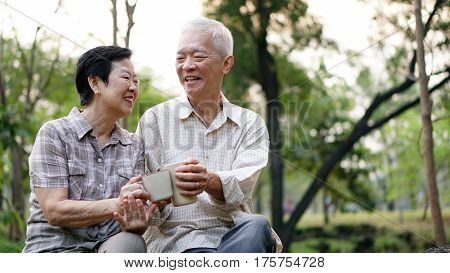 Old Asian Senior Couple Morning Date With Cup Of Coffee In Green Background Park