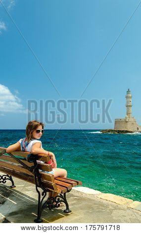 Chania, Crete, Greece - July, 13, 2013. Girl sitting on a bench in the port near the sea in front of the stone lighthouse.